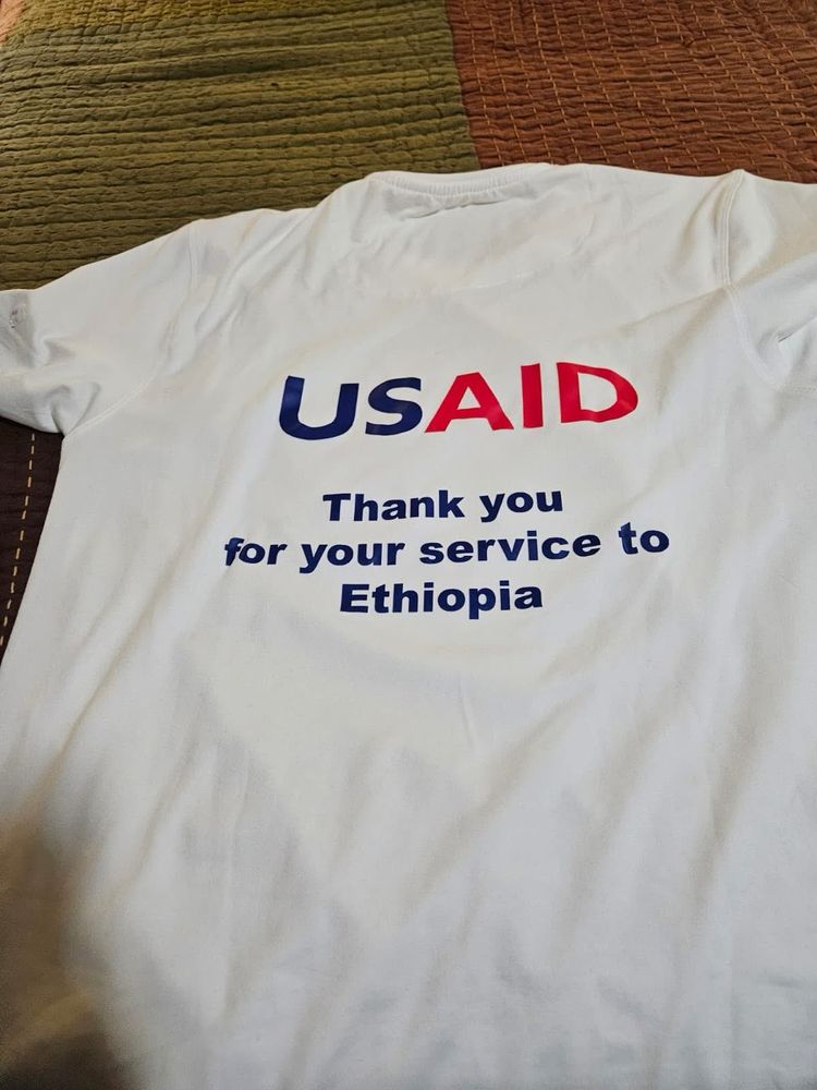 A white T-shirt is laid flat with the USAID logo printed across the chest in bold blue and red letters. Beneath it, the words read: “Thank you for your service to Ethiopia.” The shirt is simple, direct, and carries a message of gratitude.