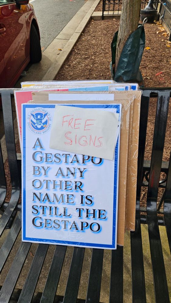 A stack of protest signs on a city bench under a tree. The top sign reads “A Gestapo by any other name is still the Gestapo,” with a handwritten note taped above saying “Free Signs.” Taken on October 18 for No Kings Day, it captures grassroots dissent reclaiming public space.