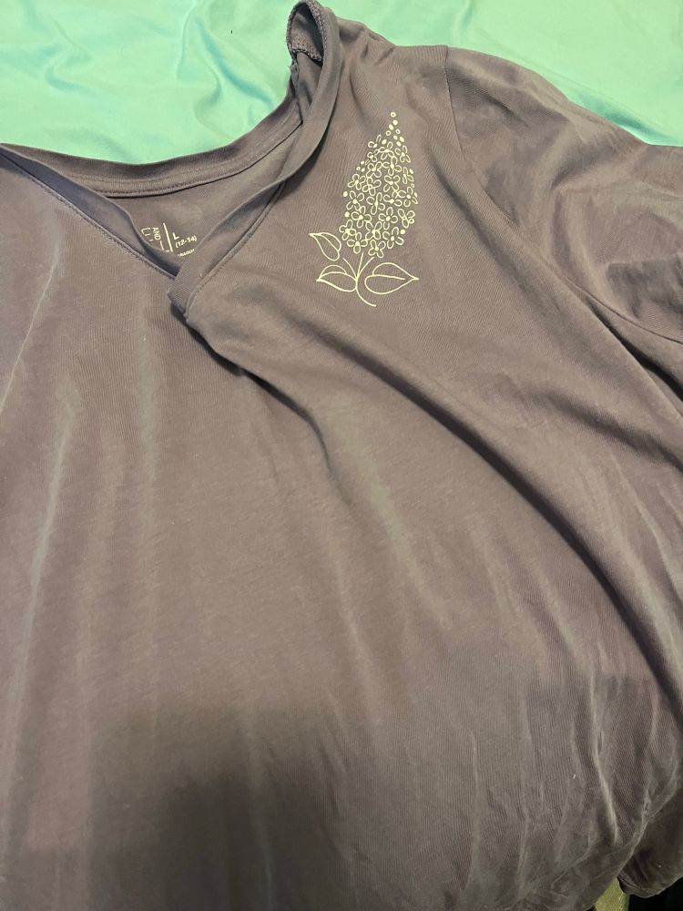 The front of a light purple shirt with a white lilac flower outline on the left shoulder 
