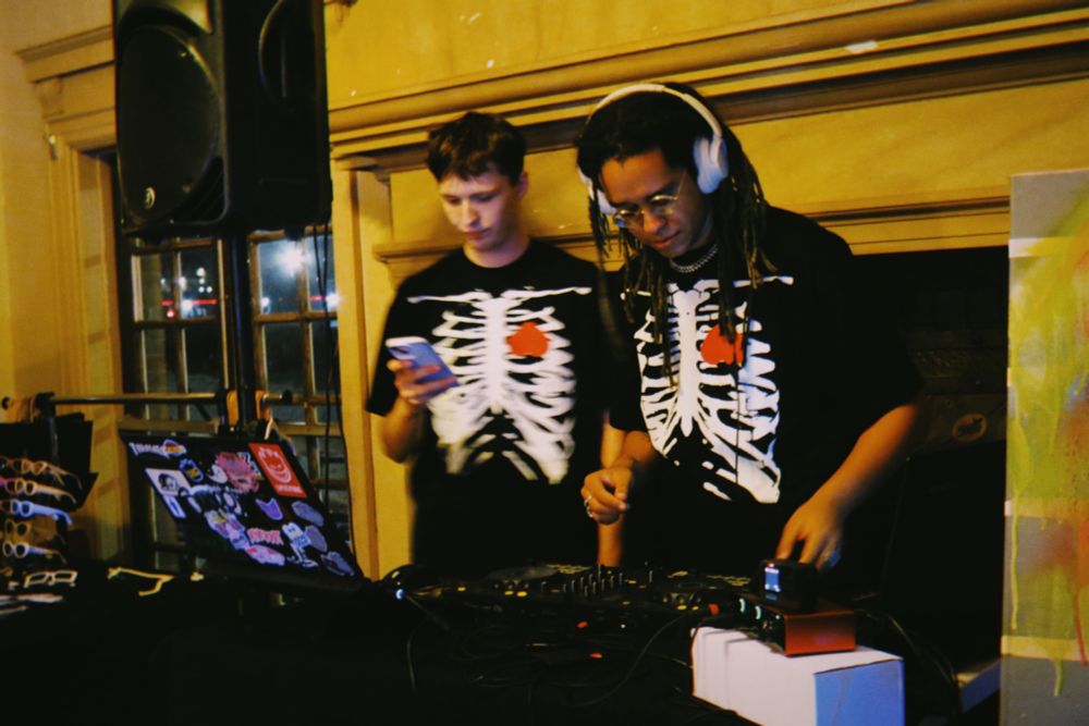 Kaash and Anki behind the dj booth, a sticker bombed laptop sits to the left while Kaash looks calculatedly at her deck. 