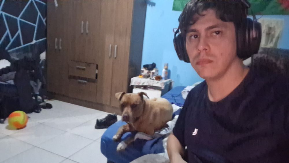 Foto de um quarto todo fudido, bagunçado, comigo em primeiro plano. Estou usando camiseta preta e headset. No segundo plano uma cadela caramelo está deitada na cama.
