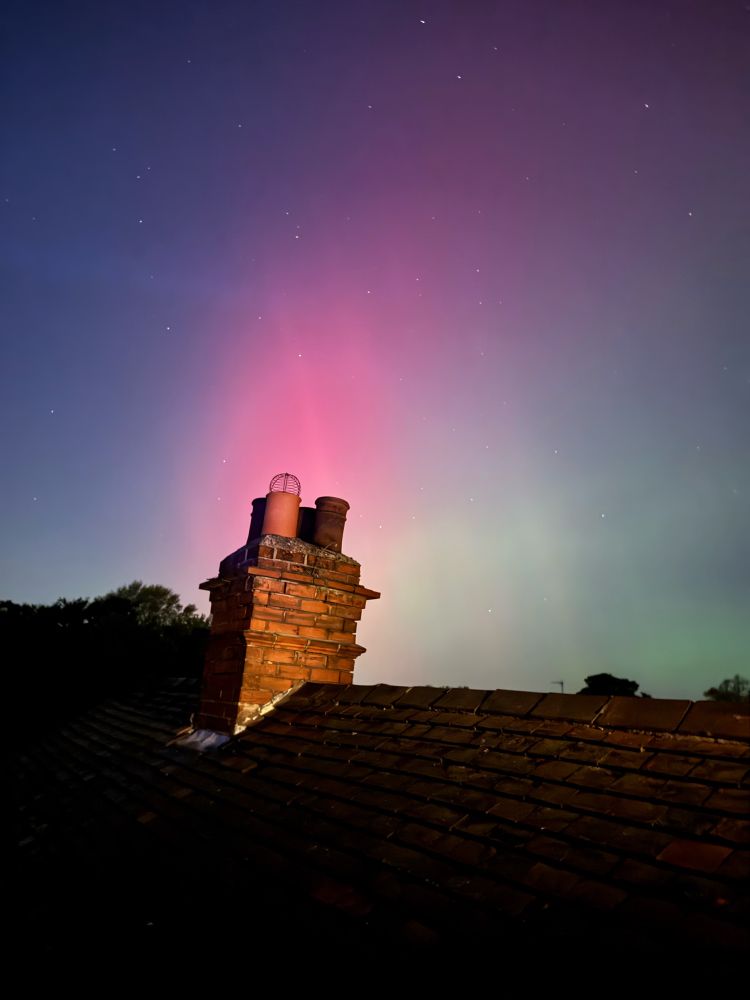 A rooftop view of the Northern Lights with a leaning chimney in the foreground backlit by the reds and greens of the aurora.