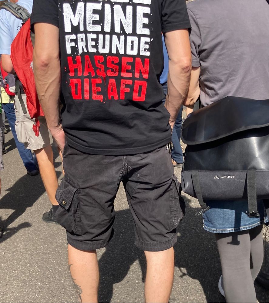 Ein Mann und eine Frau von hinten fotografiert. Der Mann trägt ein T-Shirt. Auf dem steht „alle eine Freunde hassen die AfD.“