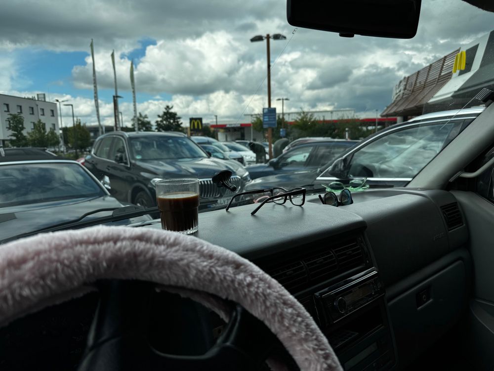Zicht vanuit de cabine van een busje op de parkeerplaats van een Mcdonald’s met een kop koffie op het dashboard. 