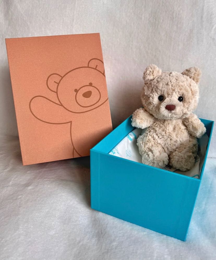 A photo of Bartholomew Bear Junior, credited to @/tamzinscosycollection on IG, where the small, beige bear plushie is sitting inside a blue box lined with seemingly tissue paper