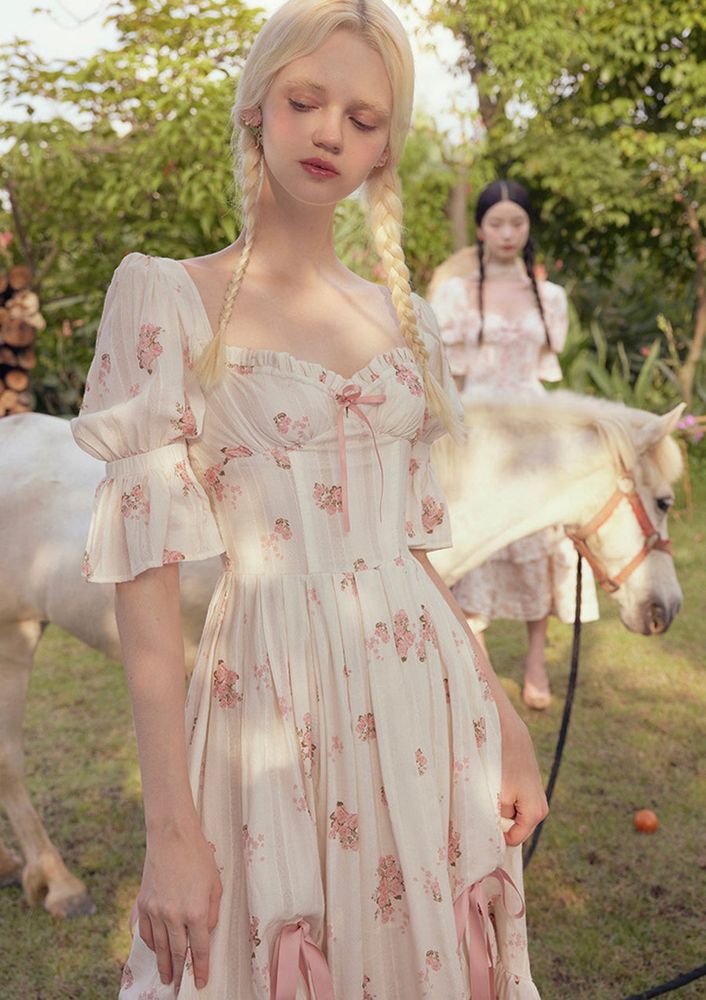 A photo of a blonde model wearing the “Rose's Smile Corset Dress Ⅱ” as listed on Lacemade for $99 USD presently; the dress is largely white or cream with a pink floral pattern and pink ribbons; not my image or my clothing, credits to Lacemade and their models 