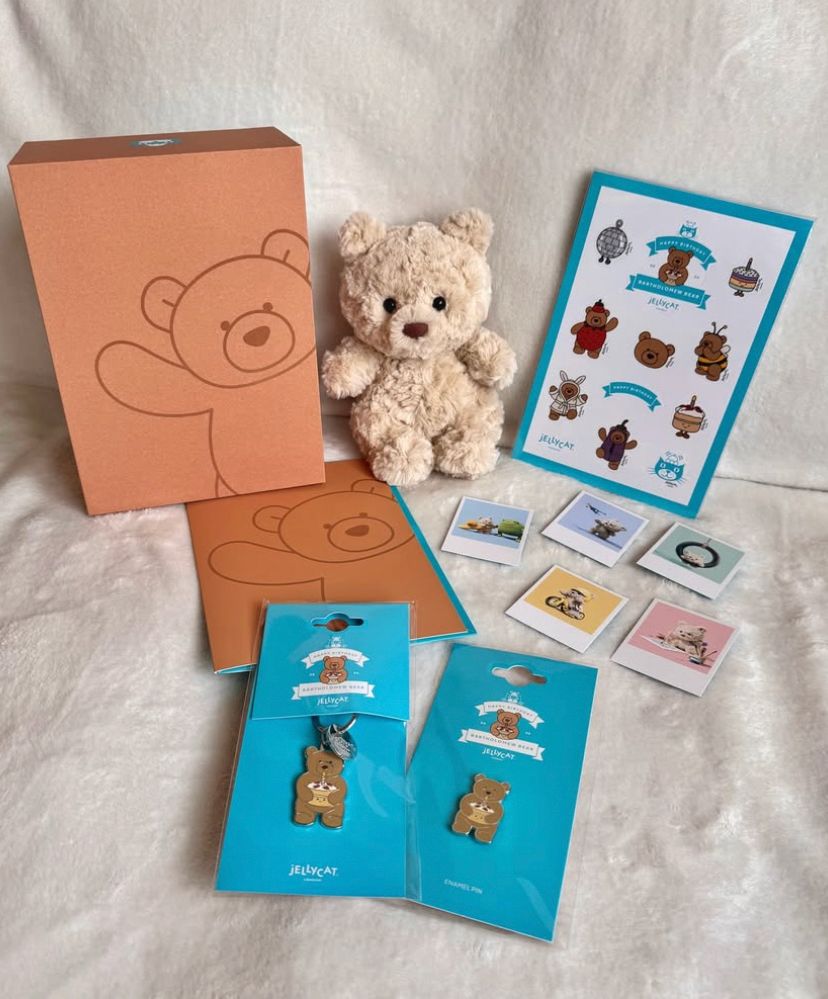 A photo of Bartholomew Bear Junior, credited to @/tamzinscosycollection on IG, where the small, beige bear plushie is sitting while surrounded by various pieces of Jellycat merch, including photos, a Bartholomew keychain, a Bartholomew pin, and Bartholomew stickers (all brown bear-themed)