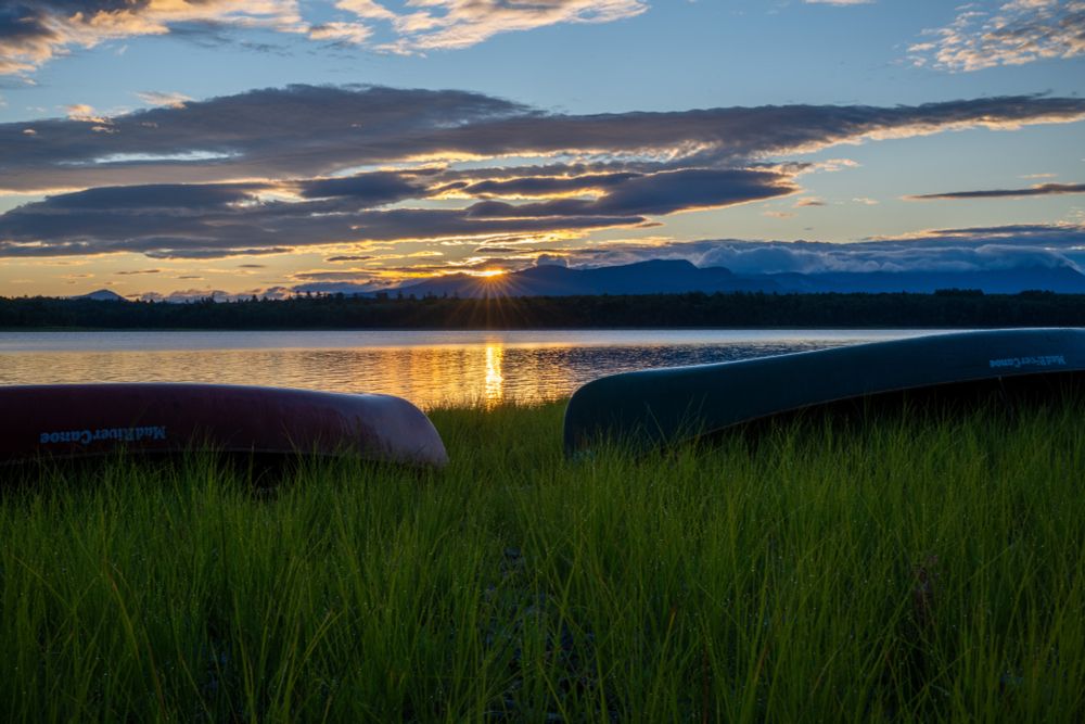 The sun rises over Mt. Katahdin.  Most of the peaks of the sprawling giant are enshrouded with clouds.  The sun's rays peak through illuminating the clouds and Lake Chesuncook.  The foreground is dominated by dew covered grass and our trusty Mad River canoes.