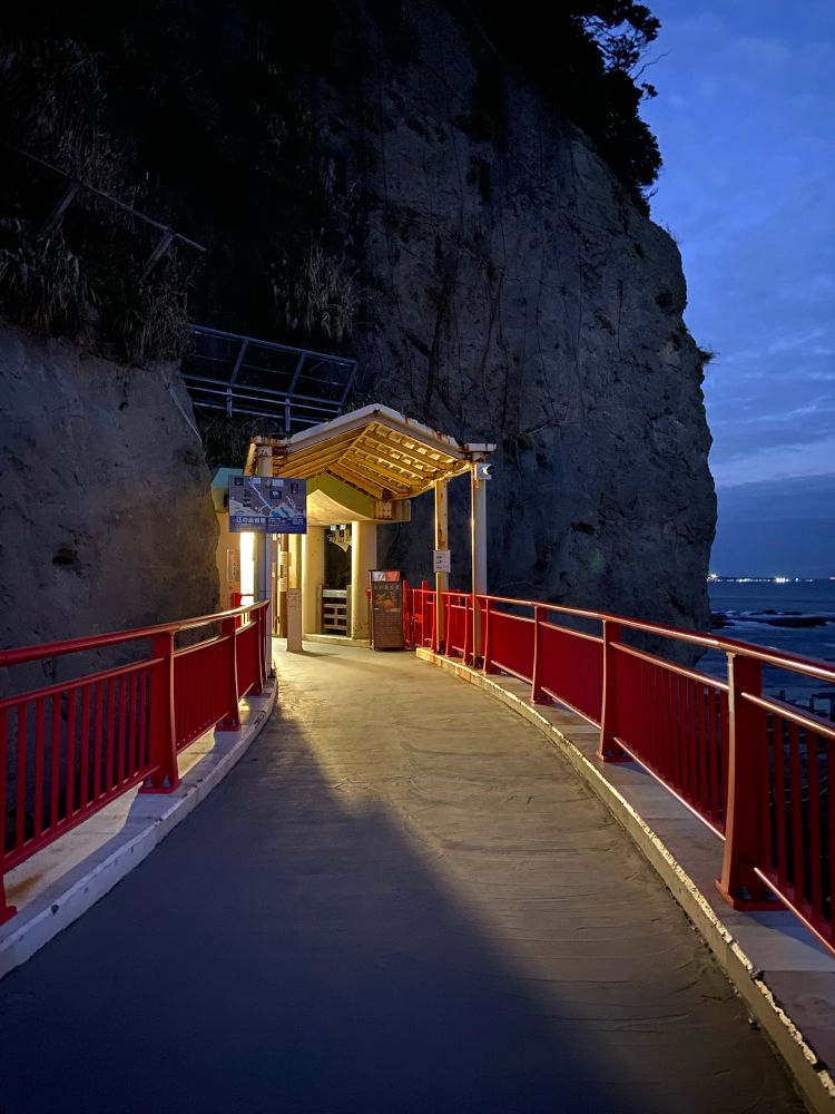 Dusk deep blue sky, red railing on a path on a cliffside overlooking the ocean, and warmly lit entrance into a cliff wall cave up ahead.