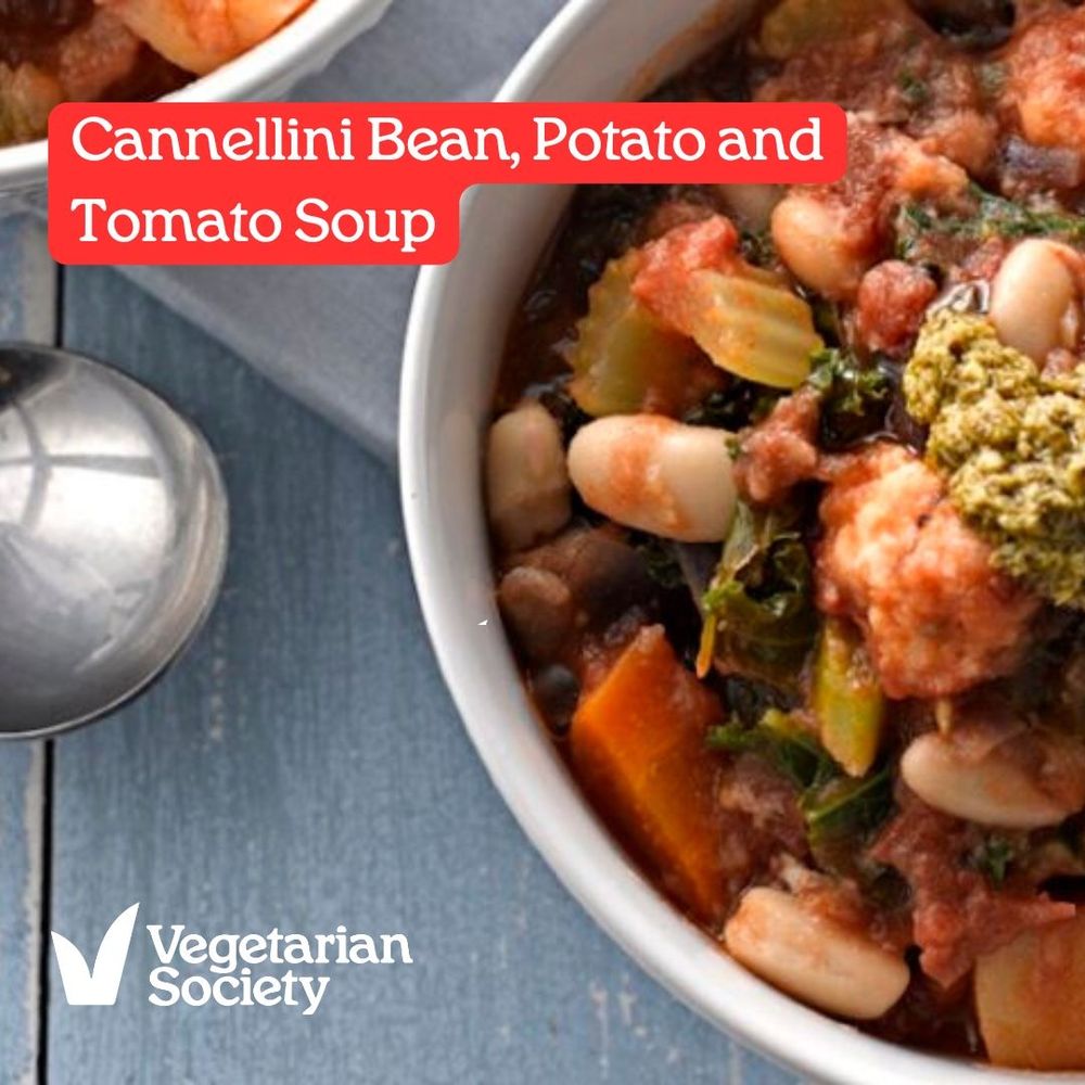 A table top with spoon and bowl. The bowl has cannellini bean, potato and tomato soup in it. 