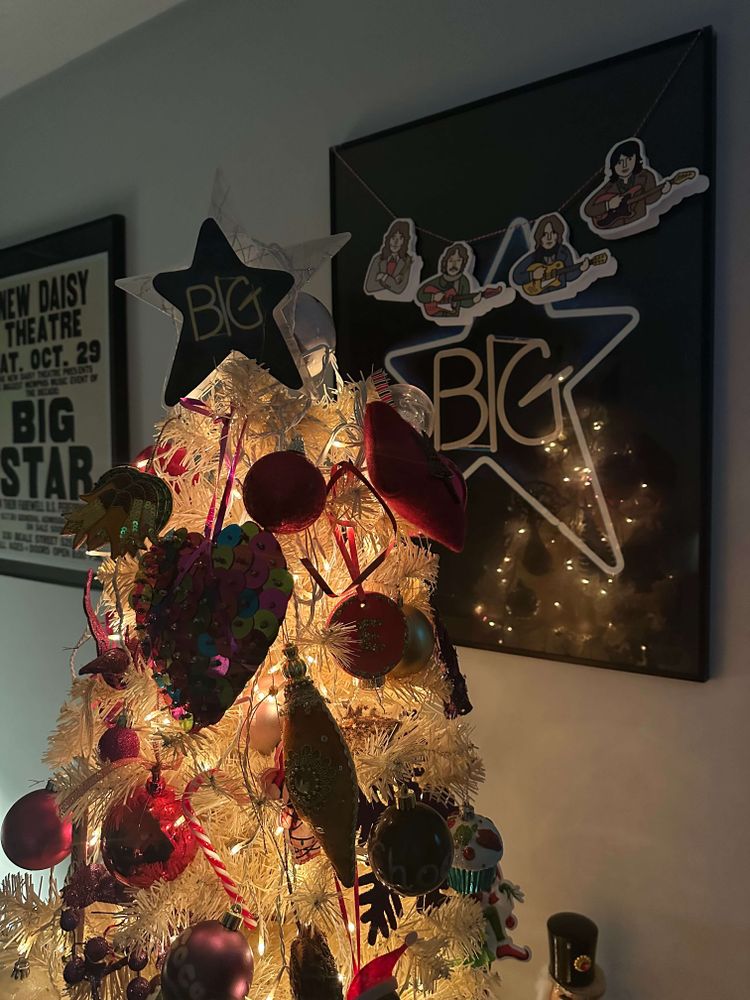 A decorated white Christmas tree adorned with colorful ornaments and topped with a Big Star, beside a framed Big Star poster, all the lads together.