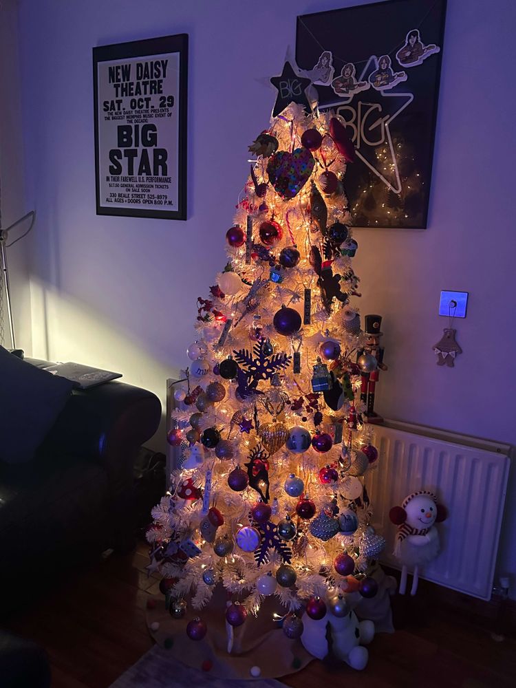 A decorated white Christmas tree adorned with colorful ornaments and topped with a star, beside some framed Big Star posters.