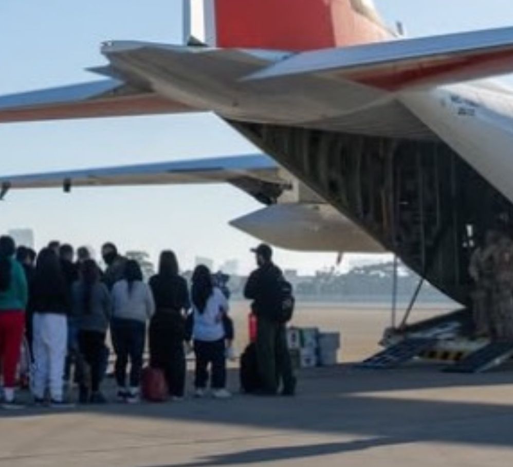 Screenshot of an official United States Coast Guard Instagram post celebrating the 100,000 flight hour milestone of their C-130J aircraft model. Featured in the celebratory photo reel is a Coast Guard C-130J parked on a ramp with 45 shackled people in the foreground waiting to be loaded on the aircraft’s cargo ramp. At the front of the line is a young woman with child in arms. 