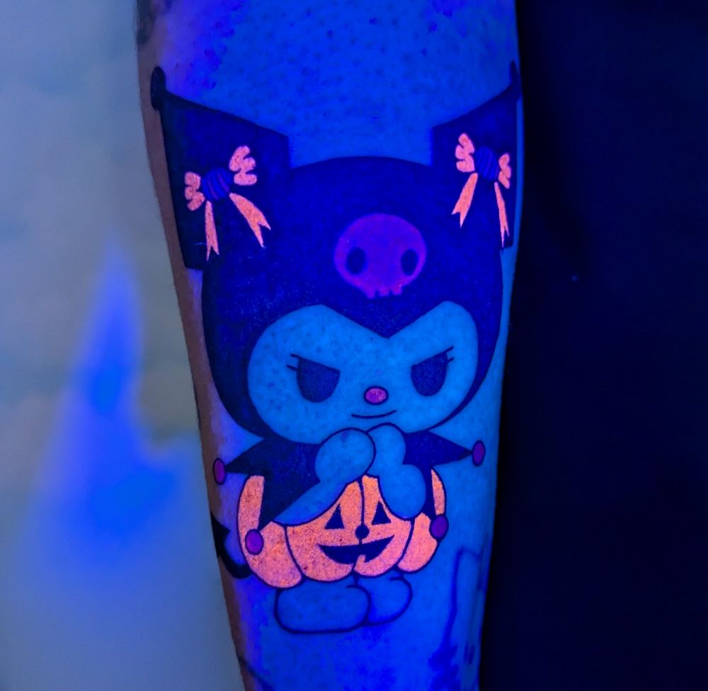 A tattoo of the sanrio character Kuromi dressed up as a pumpkin with candy bows on a forearm with UV reactive tattoo ink that make the pumpkin and bows Glow!