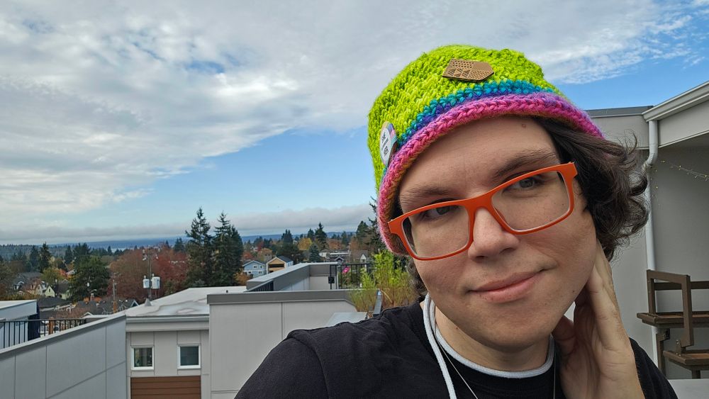 Self portrait of Teki posing on a rooftop with the Puget Sound and Olympic Mountains in the background. The sky is a partial overcast and the trademark "Blue Hole" rain shadow is clearly visible, carving a blue streak of no clouds in the sky