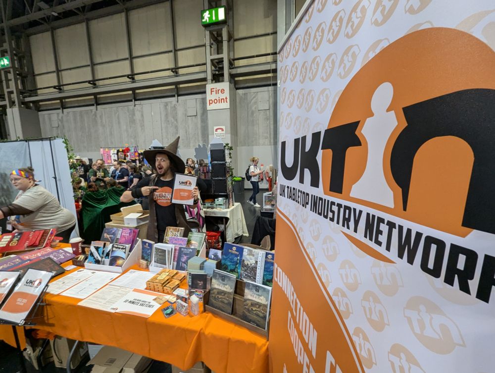 In the foreground, a banner promotes 'UKTIN'.
Beyond that is a table covered in tabletop gaming items and a busy trade hall. A person in a cool witch-style hat gestures excitedly to the stand catalogue. 