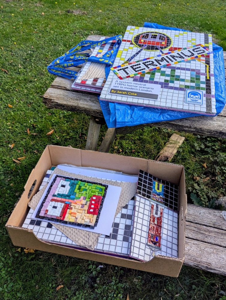 On a rotting picnic bench on a scrubby lawn, a variety of weird mosaics are stacked in a large, shallow cardboard box, and atop a couple of IKEA bags. 
