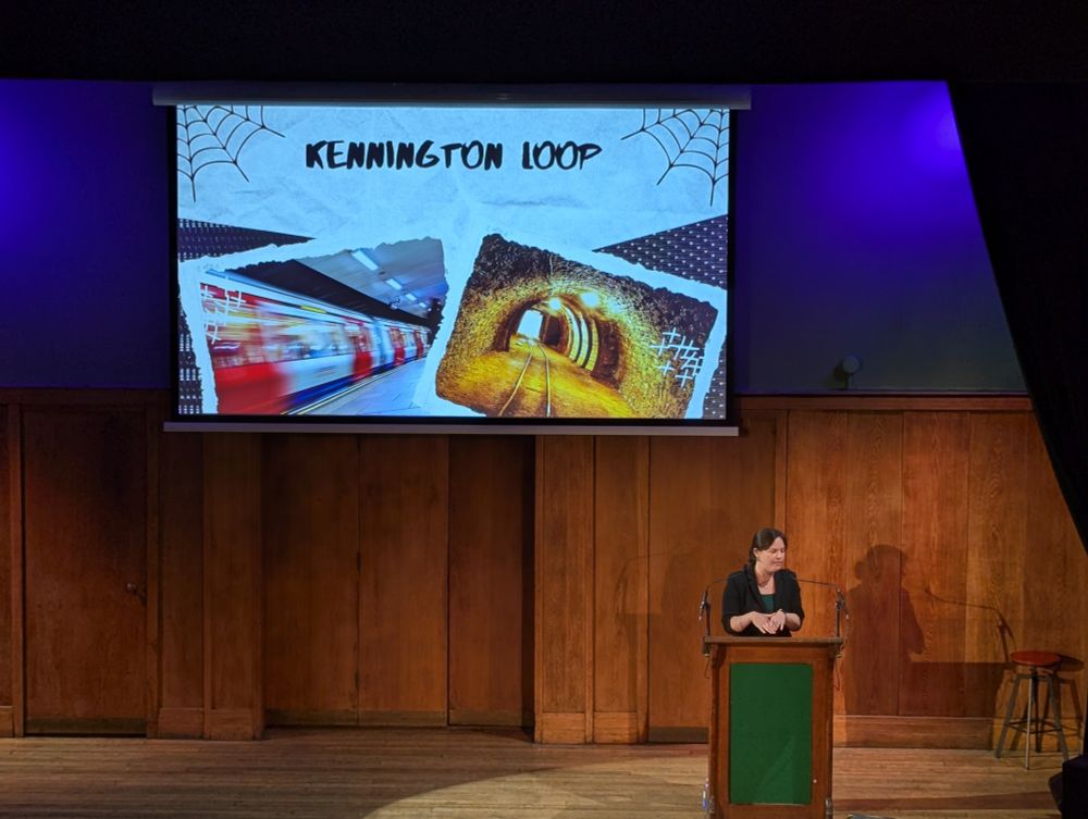 Dr E. Jay Gilbert stands on stage at Conway Hall. Behind them, the title slide of their talk says 'Kennington Loops'.