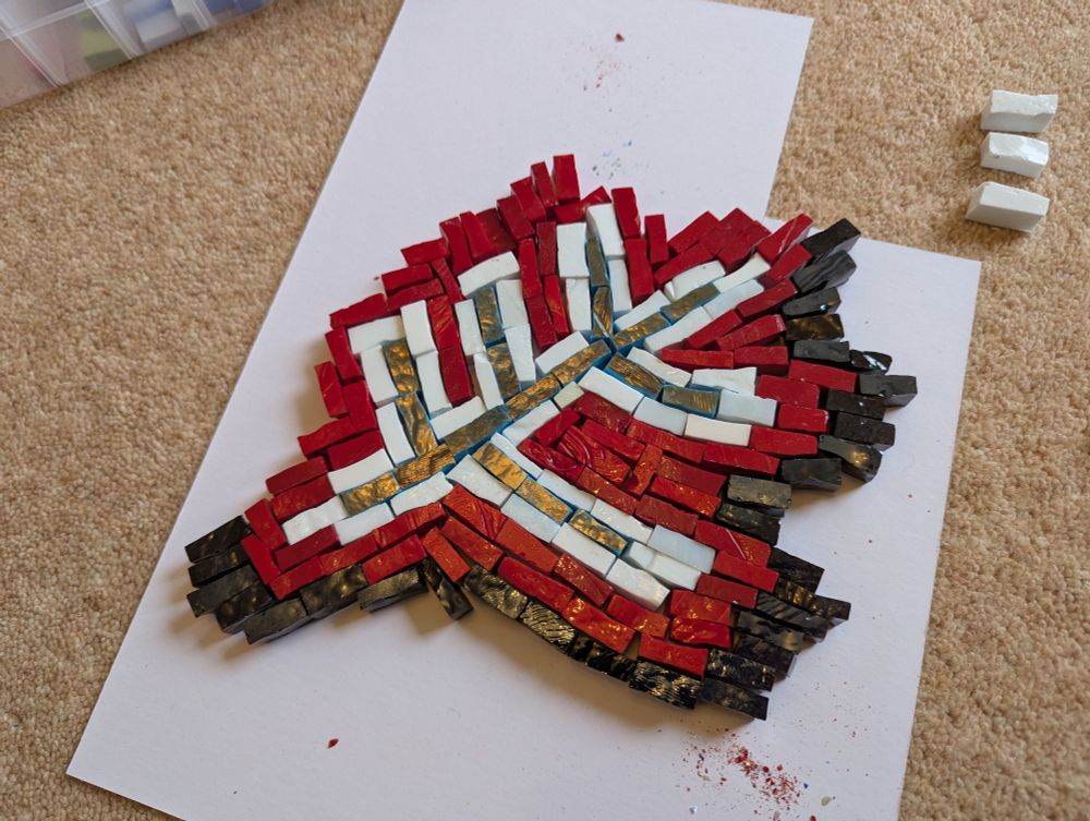 A hotchpotch and incomplete mosaic, made from irregular Smalti tiles. Black, red, and white tiles surround a vague branch shape that reflects the yellow lights of the room. This sits atop a plain white board that lies on a sandy-coloured carpet. 