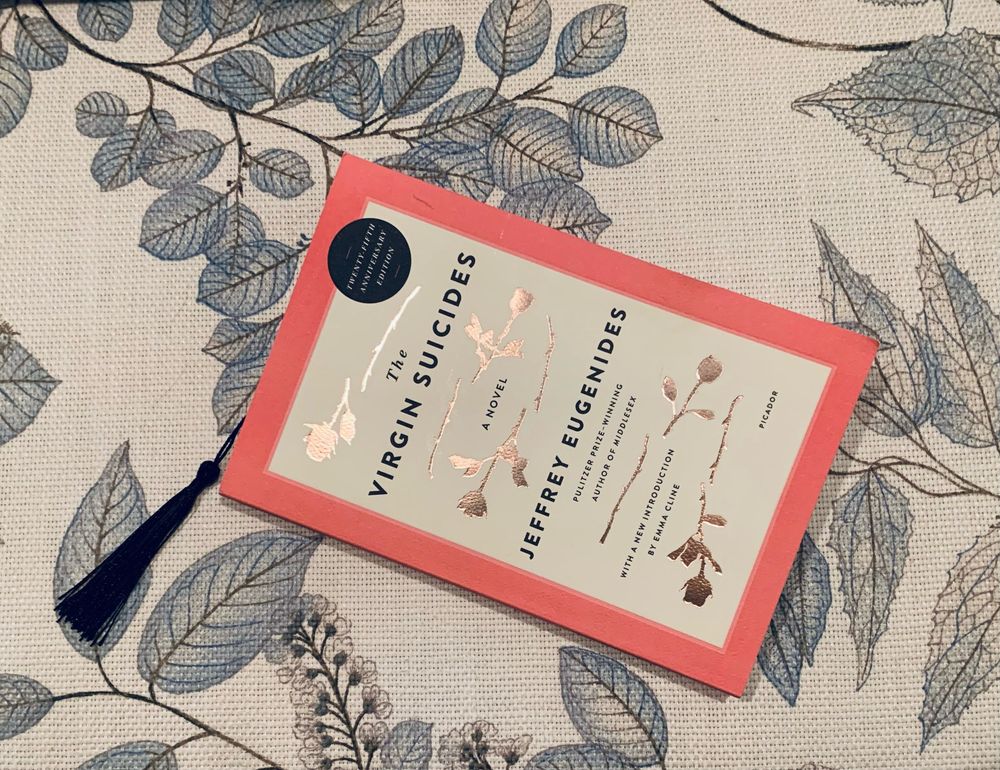 A picture of the 25th anniversary edition of The Virgin Suicides, with cover art depicting five roses with broken stems, against the backdrop of a fabric embroidered with leaves. A bookmark extends from the book. 