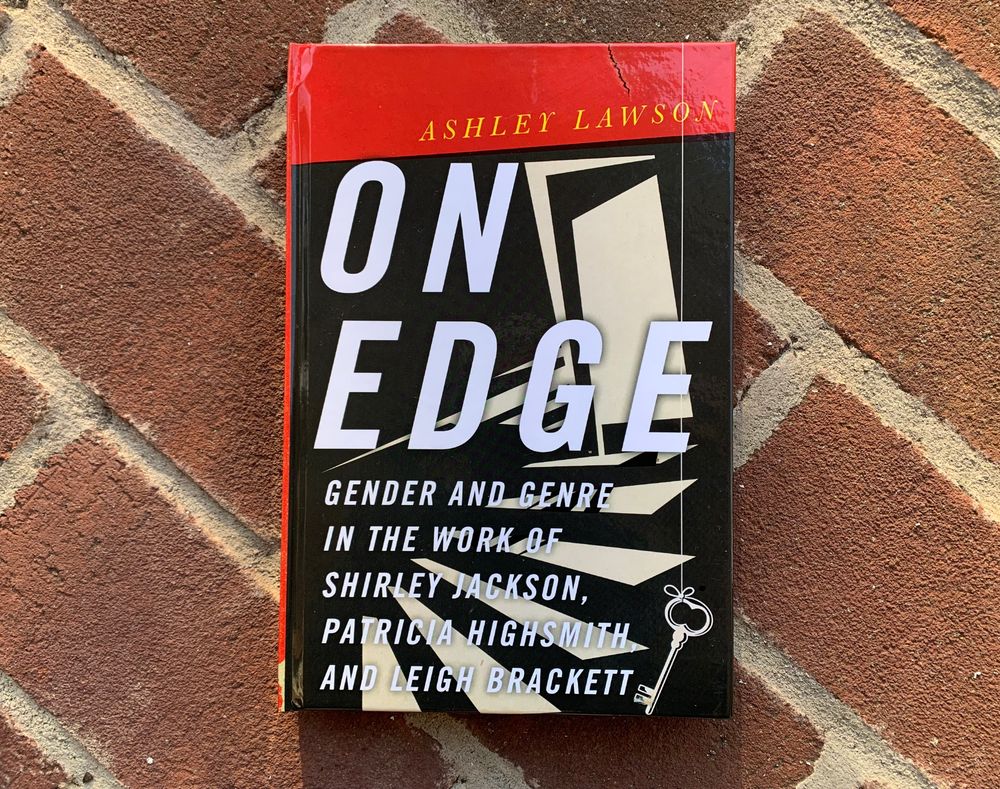 Pictured is the book On Edge: Gender and Genre in the Work of Shirley Jackson, Patricia Highsmith, and Leigh Brackett by Ashley Lawson. The black-and-white cover art shows a partially opened door leading to a shadowed staircase with a key dangling from a string in the corner.