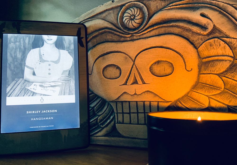 Pictured is a kindle cued up to read Shirley Jackson's Hangsaman, propped against a reproduction of New England headstone illuminated by a candle.