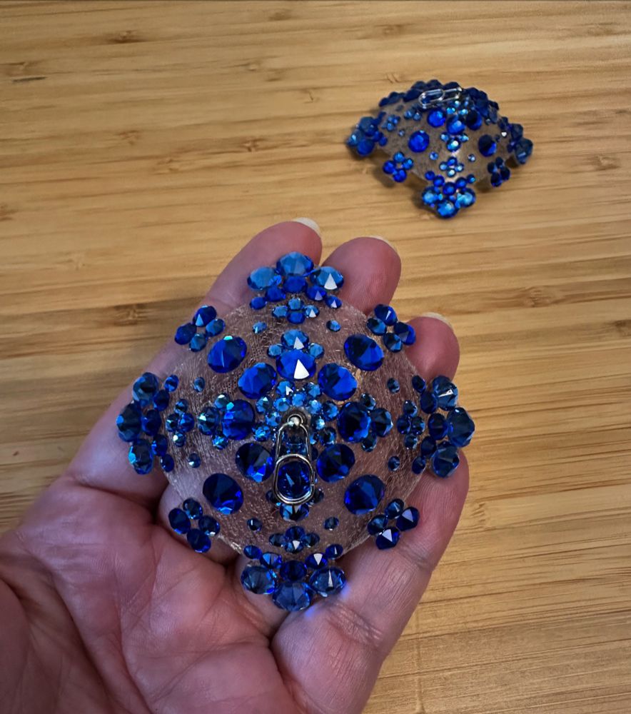 A hand holding up one of two blue crystal pasties. The other pastie is sitting on a table in the background. 
