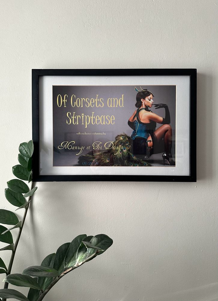 A cream coloured wall with a framed poster, showing a woman posing in burlesque attire, with a peacock tall flowing out behind her under the words “Of Corsets and Striptease, presented by Manuge et Toi Design” 