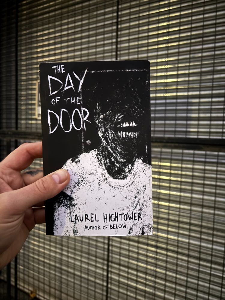 The novel, The Day of the Door by Laurel Hightower, is held up by a hand in front on a grate covered door. The black and white cover image is of a man wearing a white t-shirt with a scarred face, somewhat obscured, but with long maniacal teeth bared.