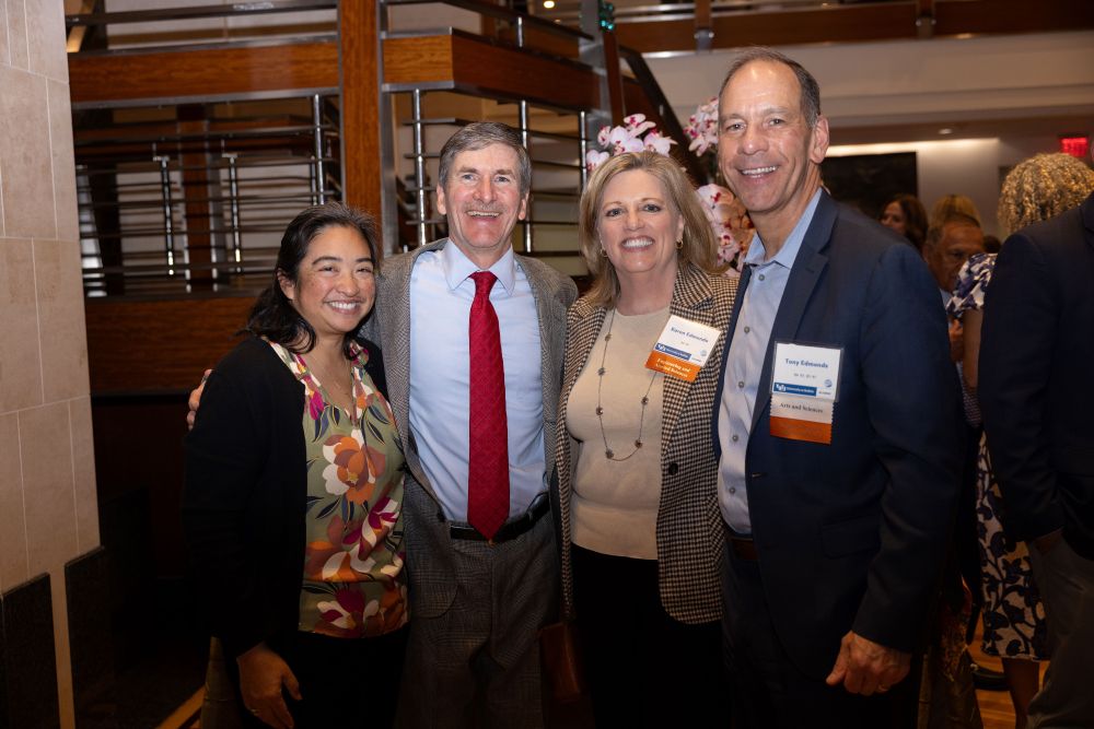 Four people stand together in the middle of a room with flowers and other people behind them.