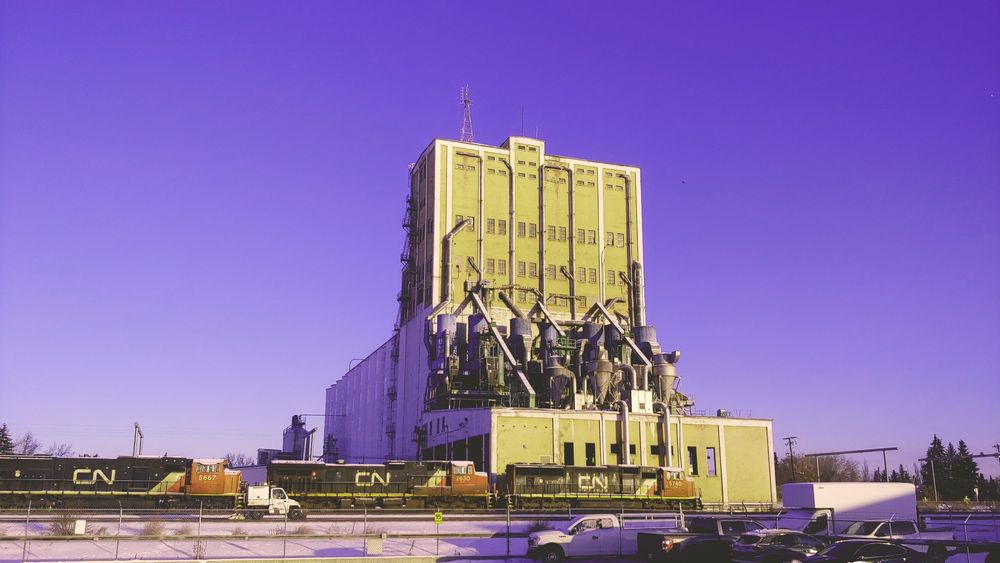 An old industrial building with tubes and loading equipment snaking over it.  It stands infront of a cold blue sky, with three train engines before it.  There is a service vehicle on a closer set of tracks, and a few visible cars in the foreground.