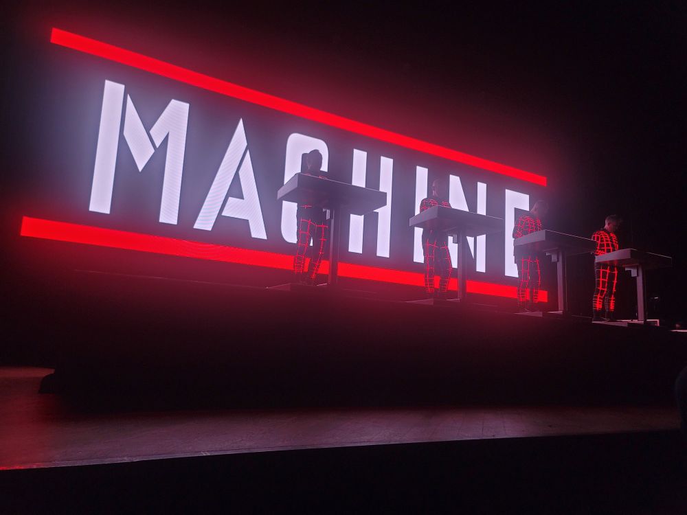 The four members of Kraftwerk stand at their little individual synth podiums on the stage at the Orpheum Theatre in New Orleans, LA. The theatre is dark, illuminated by the red LED lights on the musicians' black suits and the screen behind them, which has the word "MACHINE" in large, bright, bold white writing (and stencil font), framed by two bright red lines. 