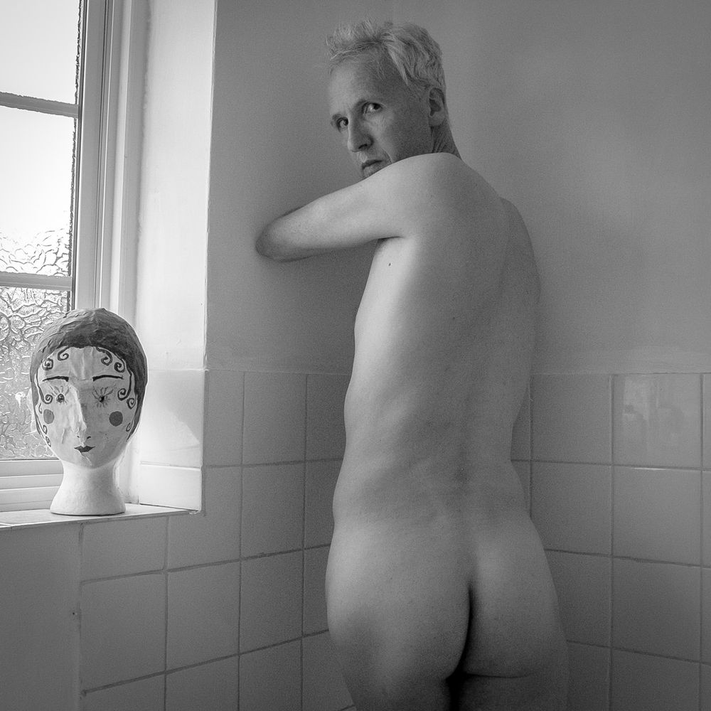 A black and white photographic portrait of the naked upper body of a man in the cornered of a bathroom. He is looking over his shoulder at the camera - on the window sill a painted polystyrene head looks on too.