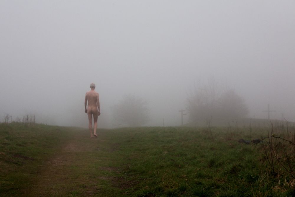 Colour photo of a naked man about 20m distant walking across a grassy field towards distant trees which are enveloped in mist.