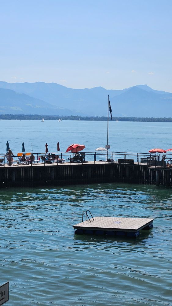 Das Seebad von Lindau, Bodensee. Man sieht einen Steg mit Sonnenschirmen, eine Schwimminsel aus Holz und im Hintergrund Berge. 