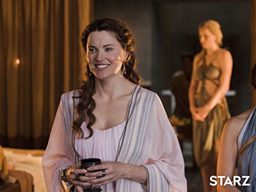 lucy lawless as lucretia batiatus in starz show spartacus:  gods of the arena. she is smiling sweetly holding a cup, dressed in a white toga with her curled, brown, mid-back length hair over her right shoulder.