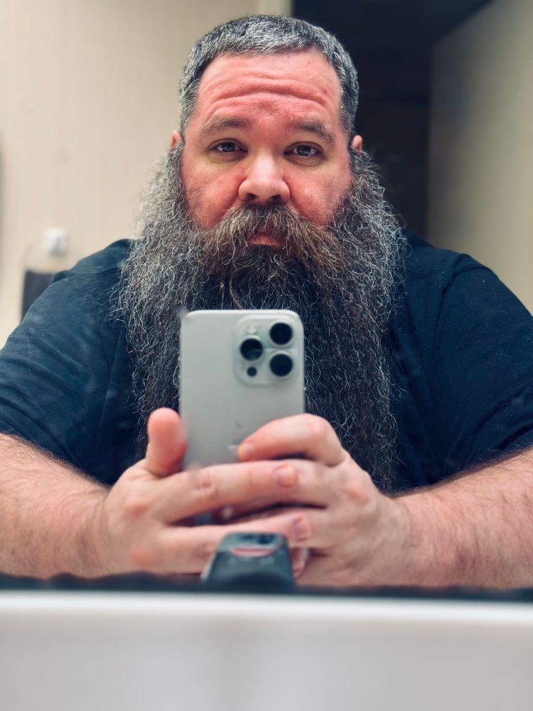 Man with long beard and salt and pepper hair taking a mirror selfie with very tired eyes. 