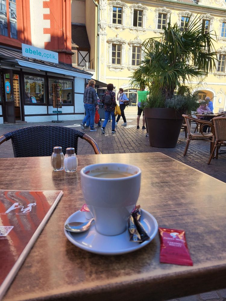 Cafe-Tisch im Freien, darauf eine Kaffee-Tasse, im Hintergrund eine Palme im Topf und ein stuckverziertes Gebäude (Falkenhaus) am Würzburger Marktplatz.