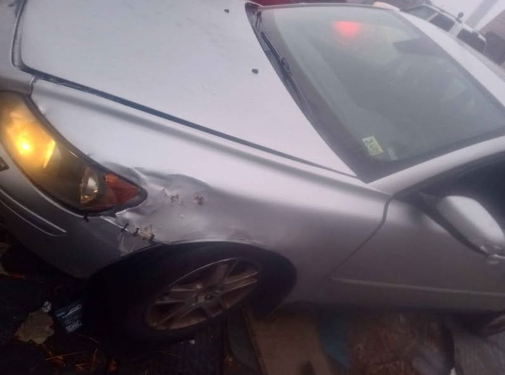 image of the same silver s40, but from the side where you can see the area around the headlight is crumpled and it’s more clear its hood is unable to close with a visible gap between it and the car. the image has the same fogginess to it 