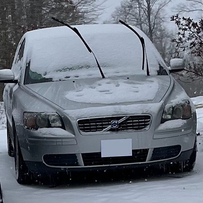 semi low quality image of a silver volvo s40 in the snow. its windshield wipers are popped up. the low beams look vaguely like eyes