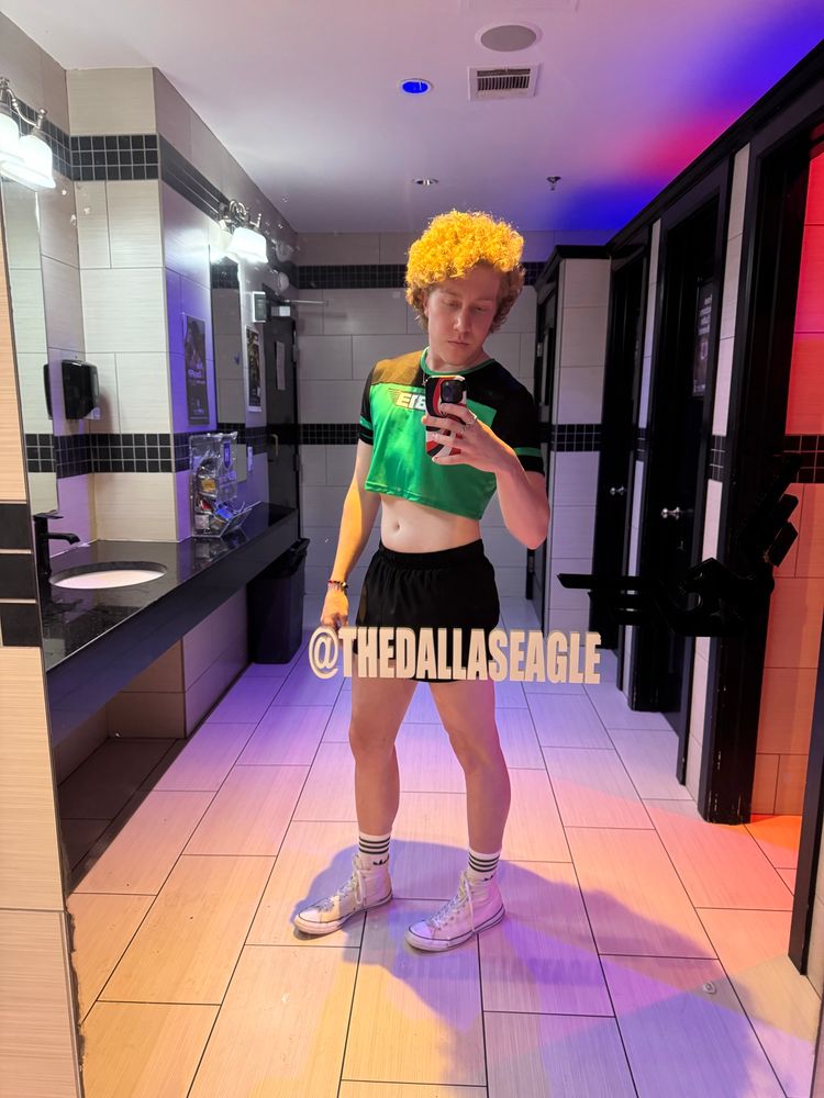 honey blond curly haired twunk wearing green and black Cell Block 13 mesh jersey crop top tee, black gym booty shorts, adidas white gym socks, and white high top Converse, taking full body selfie in bar bathroom mirror