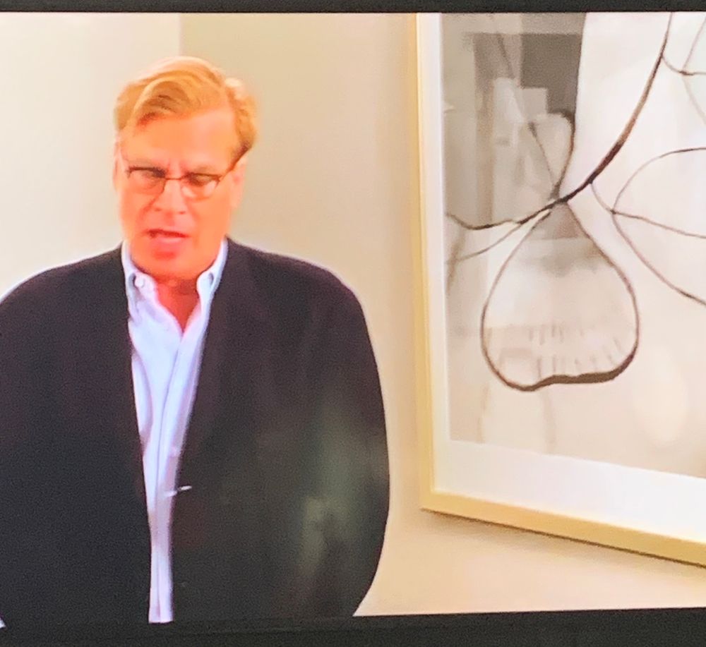 A blonde man with glasses stands next to an abstract black and white painting that looks like a set of testicles. 