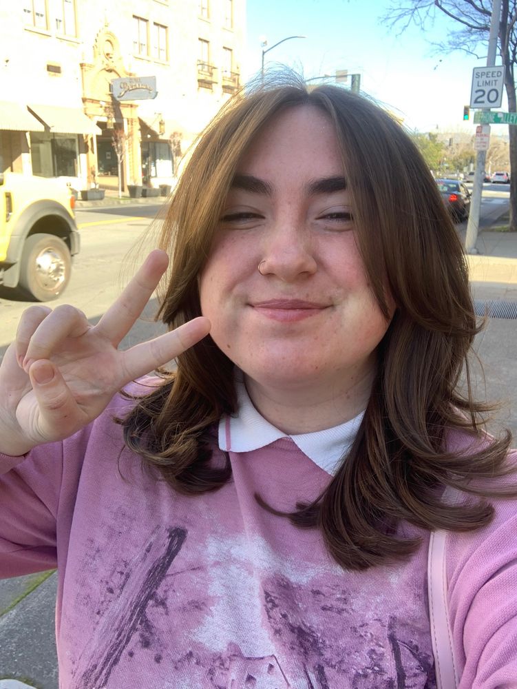 Photo of me after I got my haircut, it is shoulder length brunette hair with curtain bangs. My hand is in a peace sign