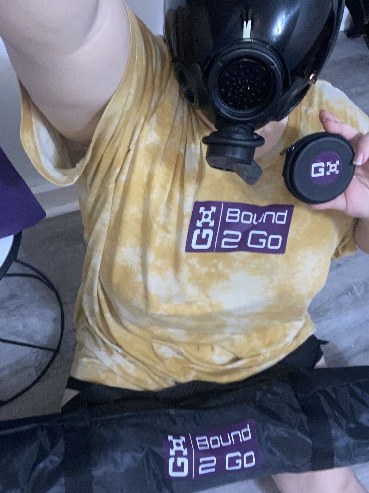 Kink drone in a MSA Millennium black gas mask, wearing a yellow shirt with the Bound2Go purple logo. It is also holding a small black zipper case with the purple Bound2Go logo in its hand and in its lap a black tent bag with the purple Bound2Go logo on it. 