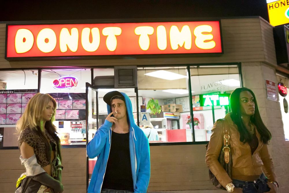 This image captures a quintessential moment from Tangerine (2015), a groundbreaking indie film directed by Sean Baker. Set on Christmas Eve in Los Angeles, the film follows two transgender sex workers, Sin-Dee and Alexandra, as they navigate the streets of Hollywood in search of truth, loyalty, and connection.

🎬 Scene Description:

• The image shows three individuals standing outside a brightly lit donut shop named Donut Time, a real location that became iconic through the film.
• The shop’s bold red and yellow signage, neon “OPEN” and “ATM” signs, and visible interior details evoke the gritty, vibrant energy of LA’s urban nightlife.
• One person smokes a cigarette while the others stand casually, suggesting a candid, unscripted moment—typical of the film’s raw, documentary-like style.
• The nighttime setting, with its artificial lighting and deep shadows, mirrors the film’s themes of marginalization and resilience.


📸 Cinematic Context:

• Tangerine was famously shot entirely on iPhones, giving it a hyper-real, intimate aesthetic.
• Donut Time serves as a central hub in the film—both a literal meeting place and a symbolic anchor for the characters’ chaotic journey.
• The image reflects the film’s commitment to portraying real lives with authenticity, humor, and heart.
