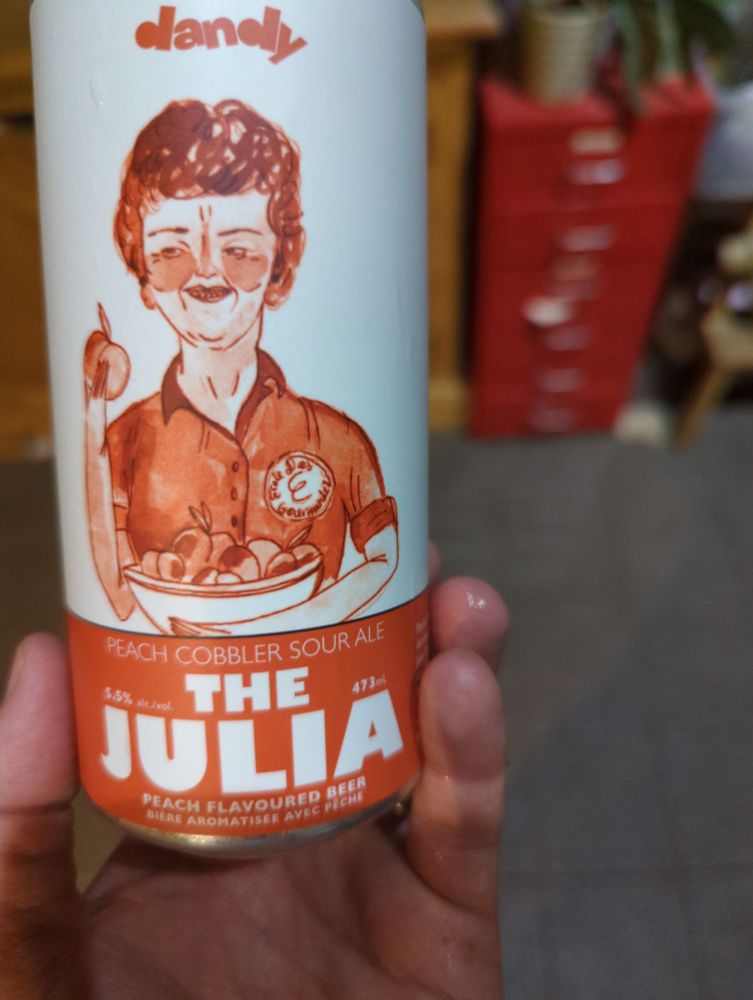 A can of Dandy Brewing's "The Julia" peach cobbler sour ale. It's a white can with an orange sketch of (I presume) Julia Child holding a peach. In the background is an out of focus tile floor and a red metal cabinet.