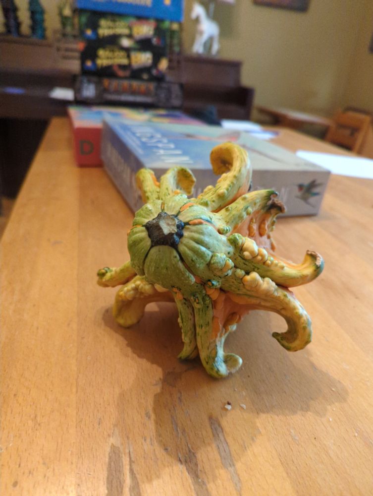 A bizarre multicolored gourd covered in ways with a strange 10 sided symmetry and a vague hint of cephalopod, on a table. There are fun board games in the background.