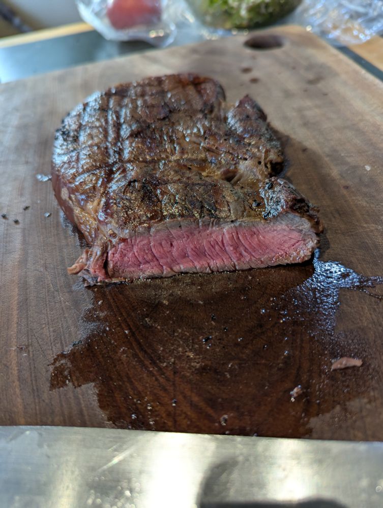 Grilled sirloin steak, nicely medium rare, cute open on a dark wood cutting board. Some chimichurri is barely visible in the background