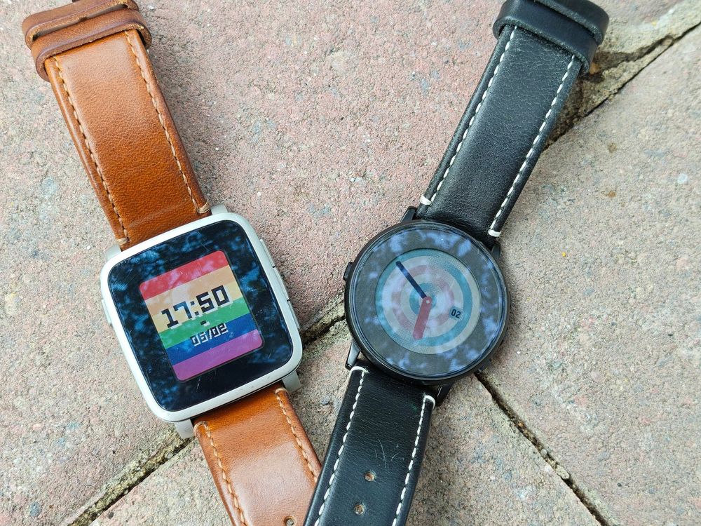 Two Pebble Time smartwatches side by side on a grassy backdrop. The round watch to the left shows an analog clock face with concentric rings in transgender pride colors, and the rectangular watch to the right shows a digital clock face backed by a six-stripe rainbow flag.