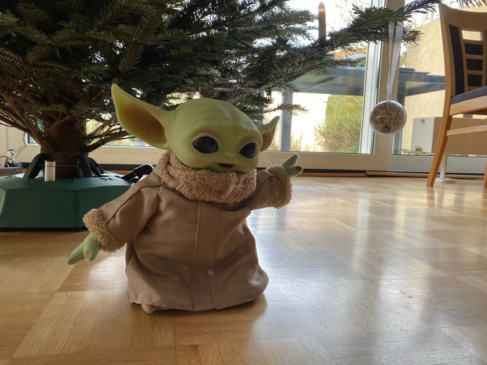 Grogu aus Star Wars hebt die linke Hand und lässt mit Hilfe der Macht eine Christbaumkugel schweben.